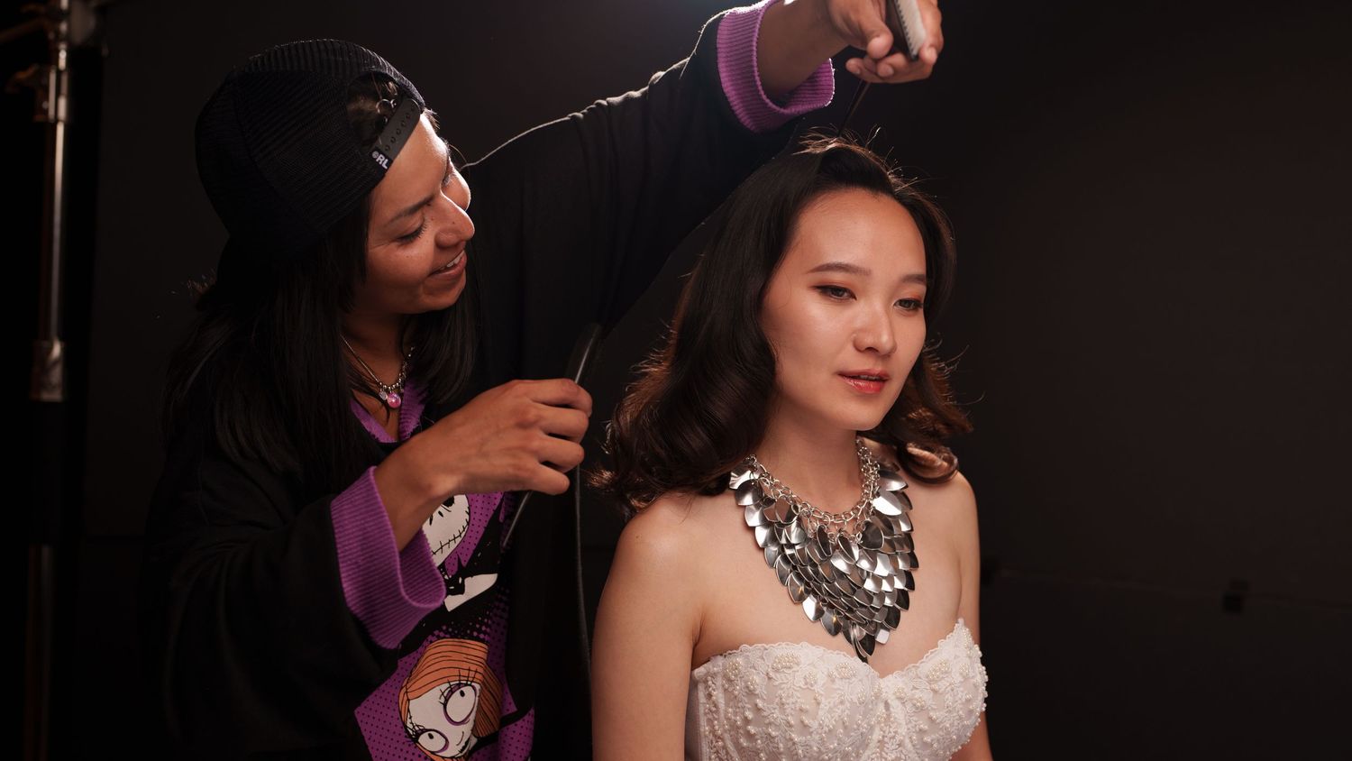 A makeup artist styling a young Asian woman in a white dress with a statement necklace in a studio setting.