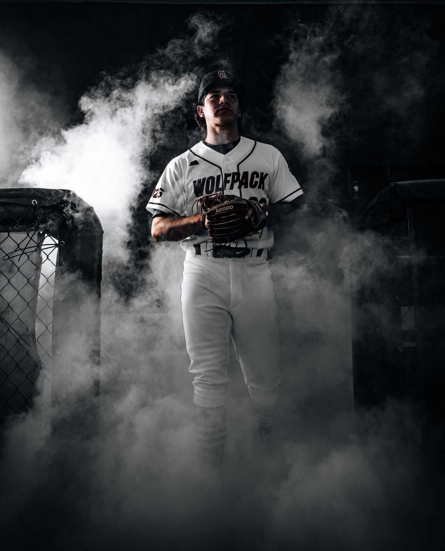 Baseball player in white uniform stands dramatically in atmospheric smoke during professional sports photo shoot.