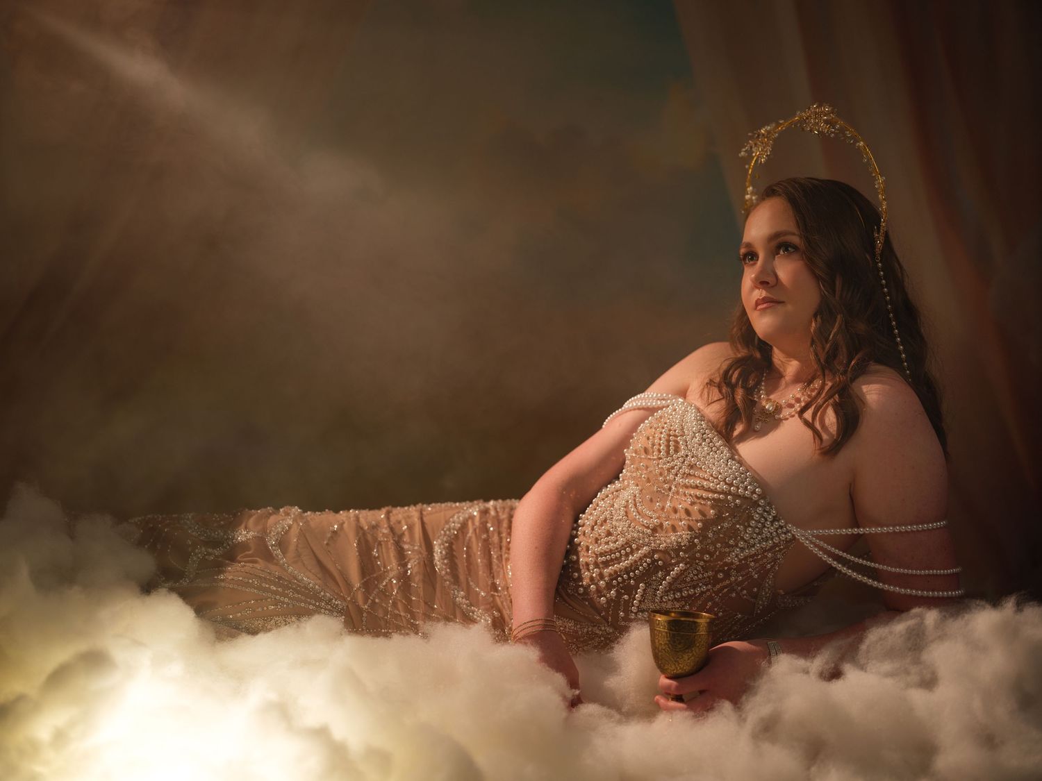 Woman in beaded gown and gold crown reclines on clouds holding a goblet in a dramatic, ethereal studio portrait.