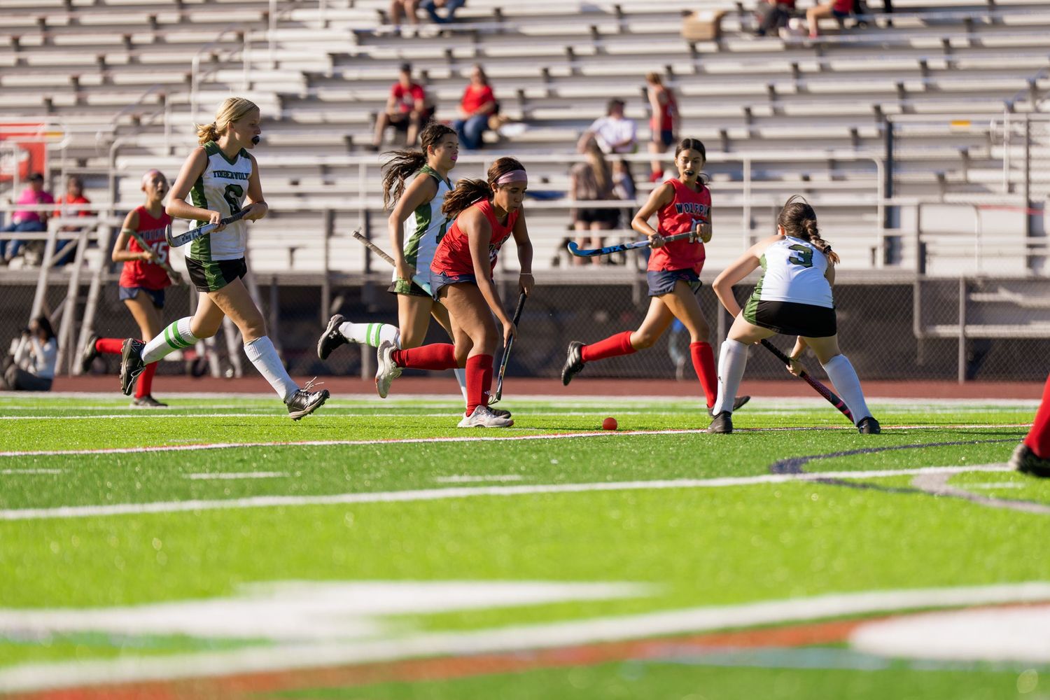 Soccer players compete on a turf field during an intense match with spectators watching from stadium bleachers.