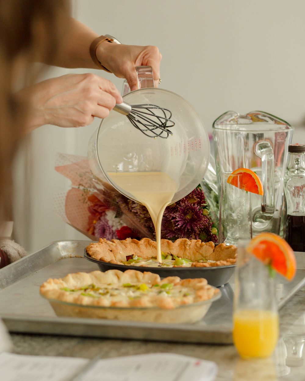 Woman pouring custard mixture into a pie crust on a baking tray with mimosas and flowers in background.