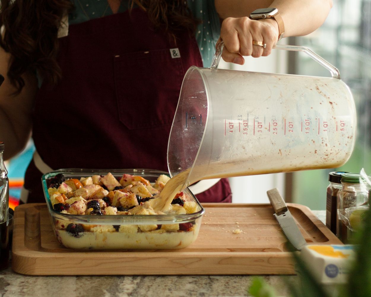 Person pouring liquid from a large measuring cup into a glass baking dish with bread pudding on a wooden board.