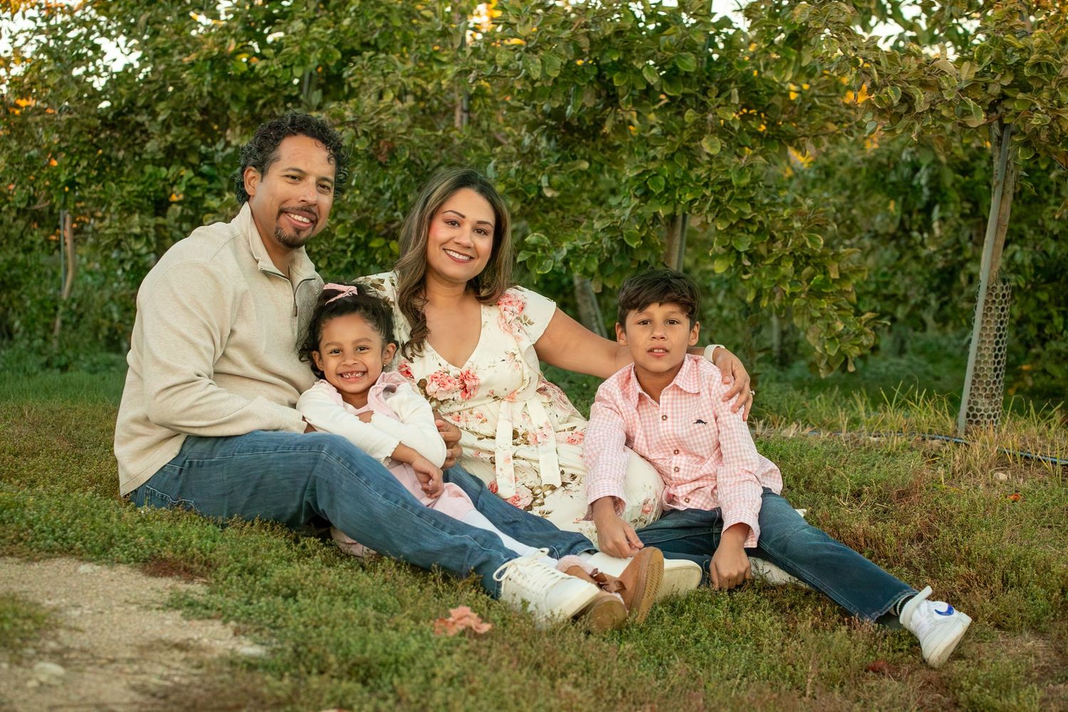 A family of four sits together on grass near an apple orchard on a sunny autumn day.