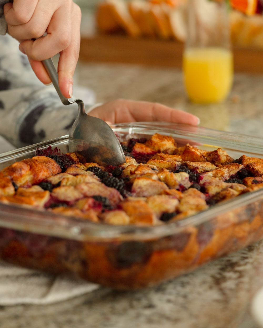 Person scooping berry bread pudding casserole from glass baking dish with orange juice in background.