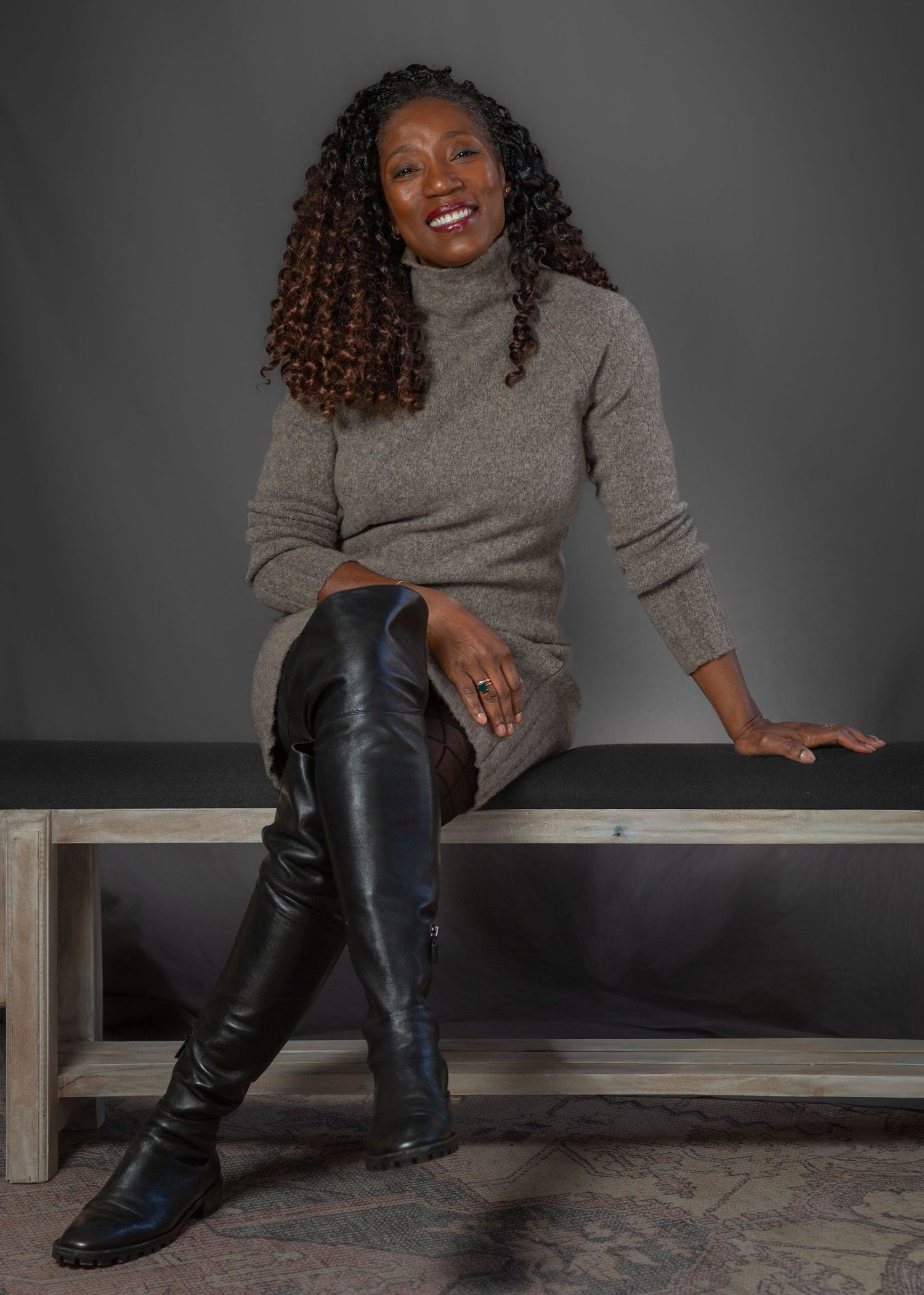 Woman in gray sweater and leather pants poses casually on wooden bench in artistic photo series.