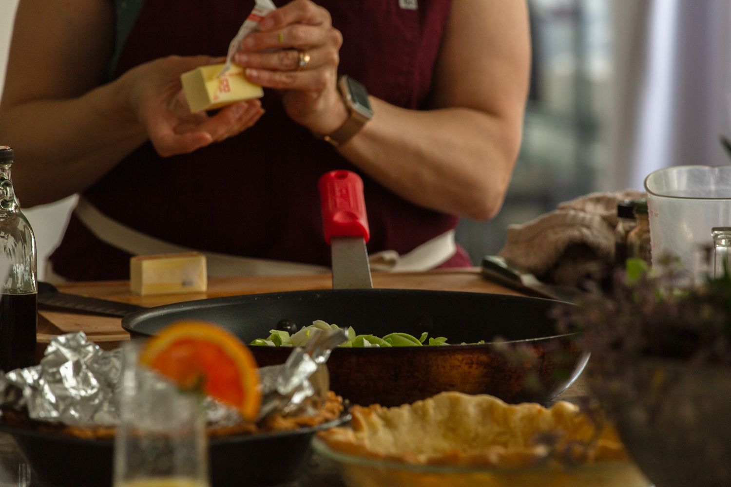 Person grating cheese over a skillet of vegetables on a wooden cutting board with baked goods nearby.