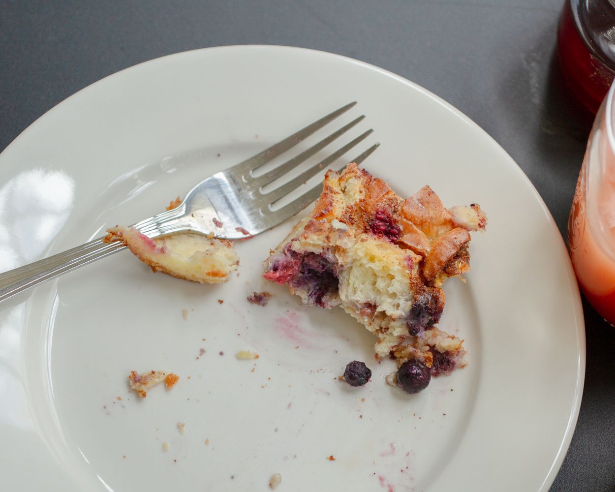 A white plate with a partially eaten blueberry pastry slice and a silver fork, with crumbs scattered around.