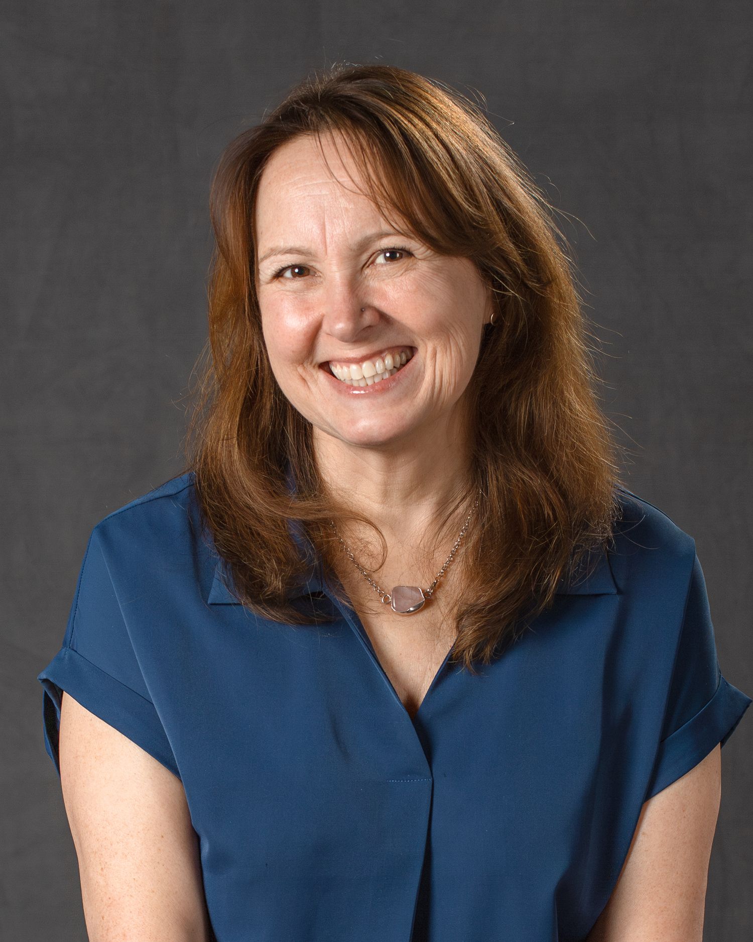 Smiling woman with auburn hair wearing blue blouse and necklace in professional headshot photo.