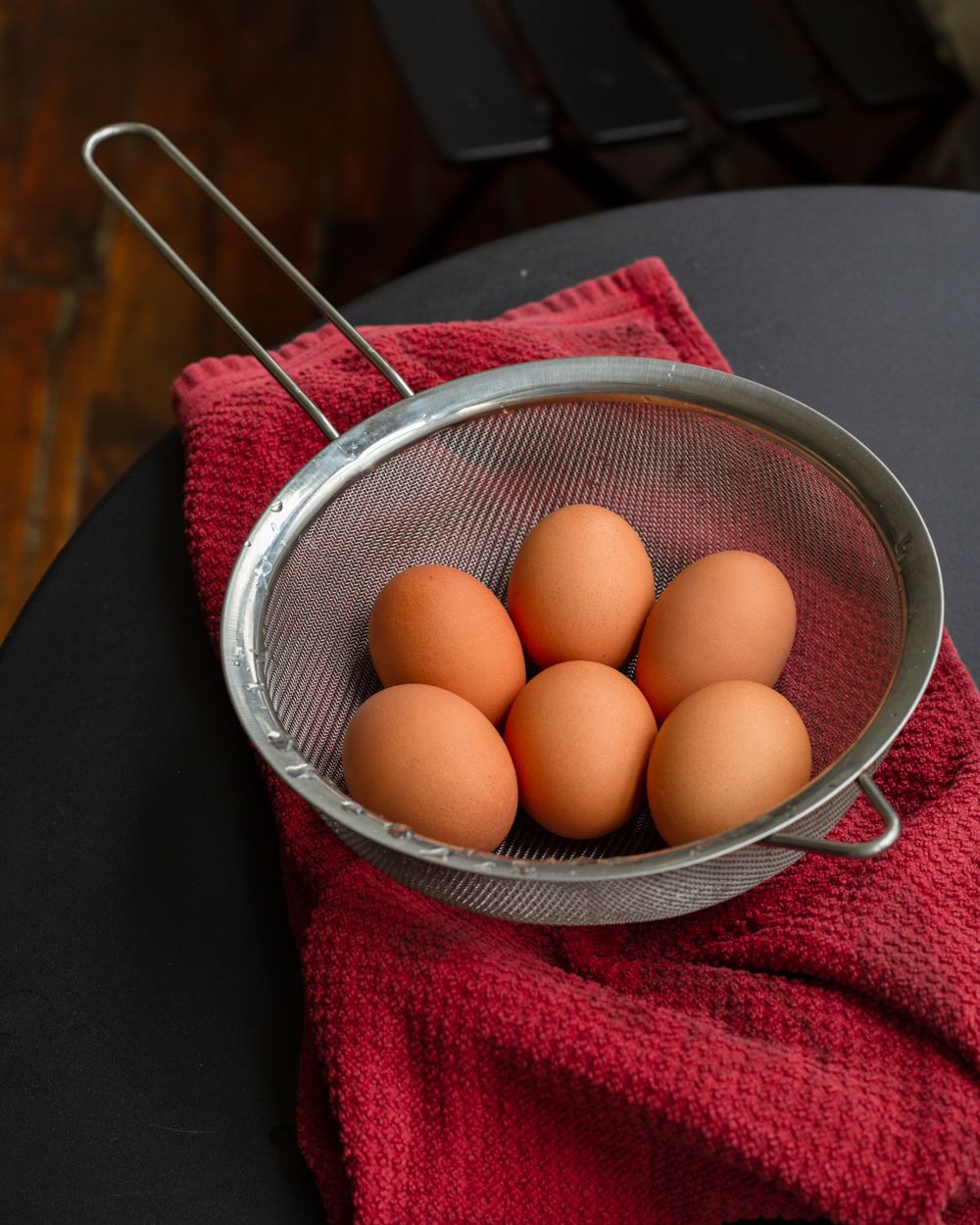 Six brown eggs resting in a metal mesh strainer on a red cloth napkin on a dark round table.