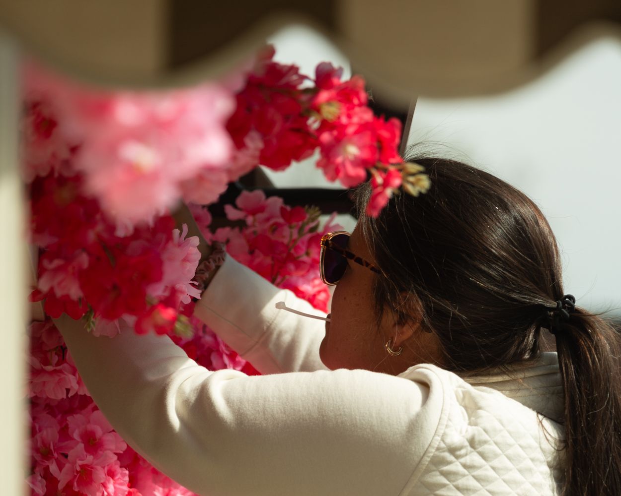 Woman in white jacket arranging vibrant pink and red artificial flowers outdoors in bright sunlight.