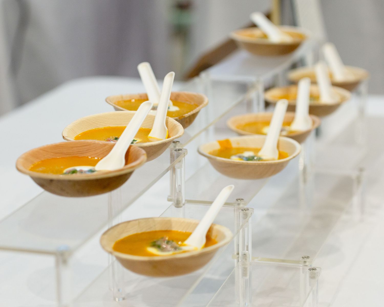 Rows of elegant butternut squash soup in ceramic bowls with white spoons displayed on acrylic risers at a catered event.