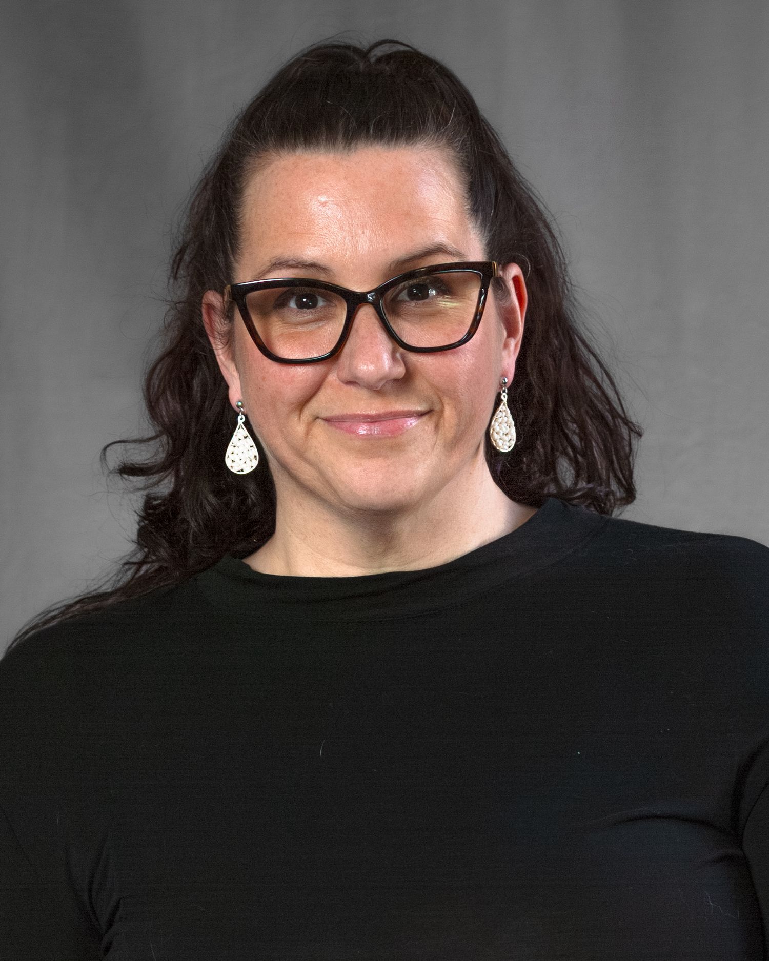 Professional headshot of someone wearing black-rimmed glasses and a black top with teardrop earrings against a gray background.