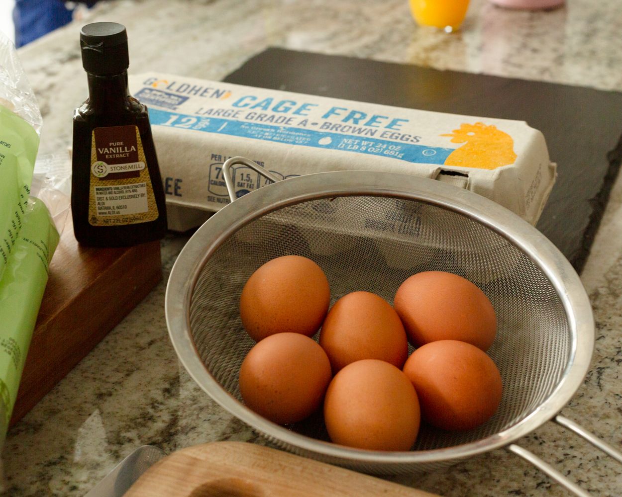 Cage-free brown eggs in a mesh strainer beside vanilla extract on a granite countertop.