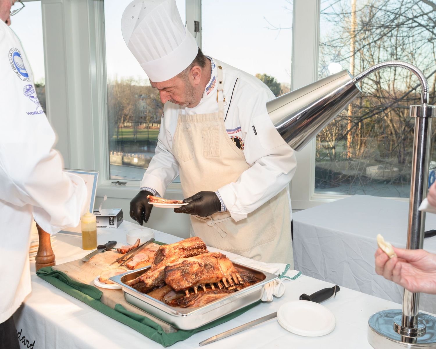Chef in white uniform and hat carving roasted meat on a tray at an upscale catering event near large windows.