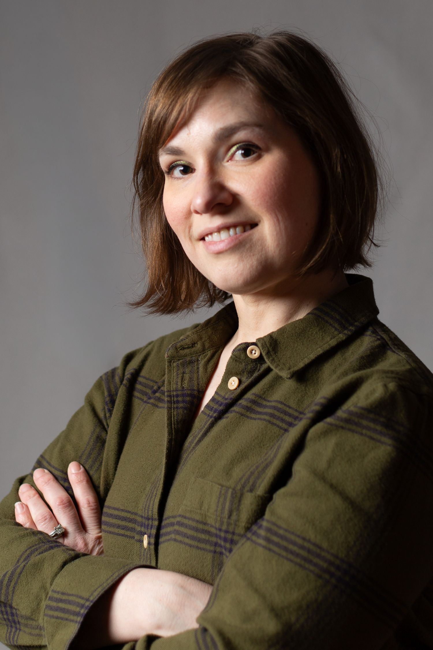 Professional studio portrait of a person in an olive green plaid shirt with crossed arms and a warm smile.