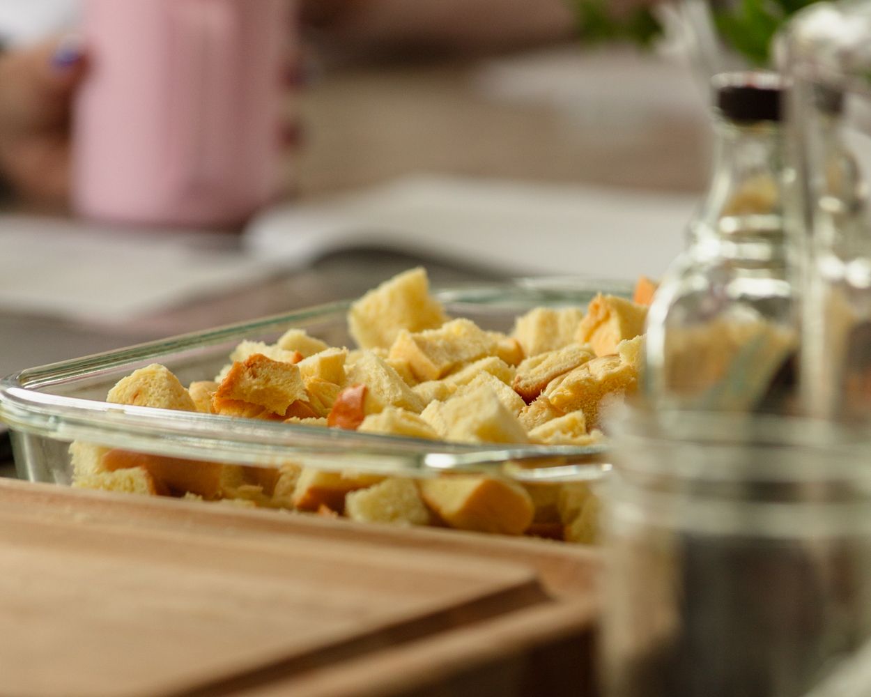 Glass baking dish filled with golden croutons on a wooden cutting board beside glass condiment bottles.
