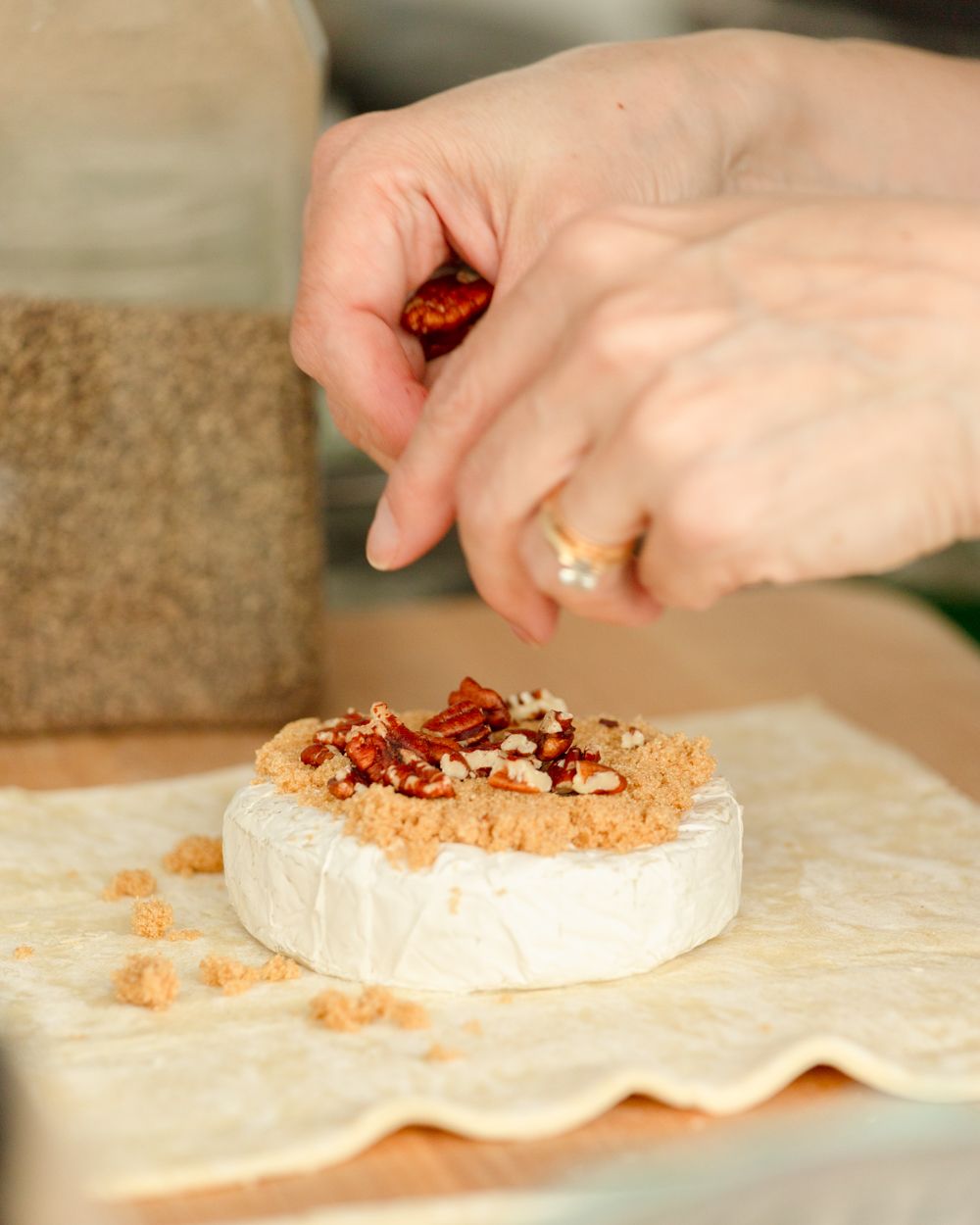 Hands placing pecans on brie cheese wheel wrapped in pastry dough on parchment paper.