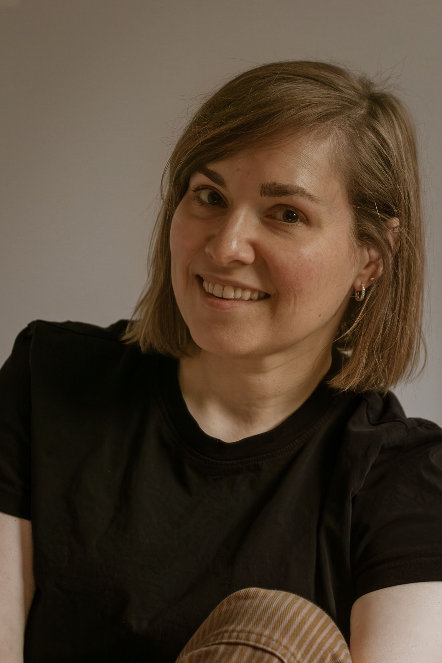 A person with shoulder-length brown hair wearing a black t-shirt smiles warmly against a neutral background.