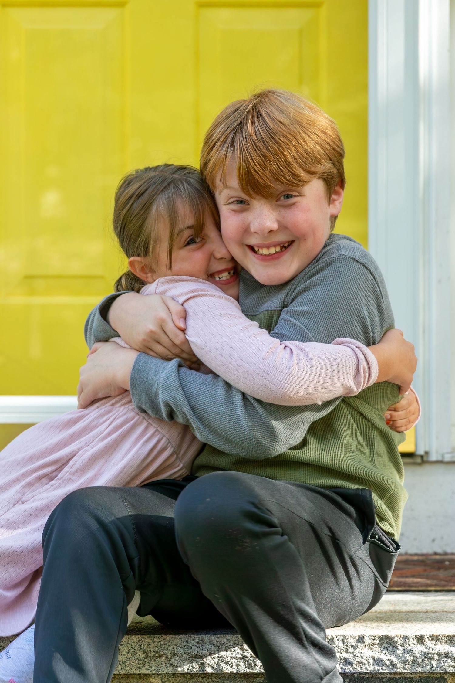 Two children in casual clothing share a warm embrace while sitting together on a porch step.