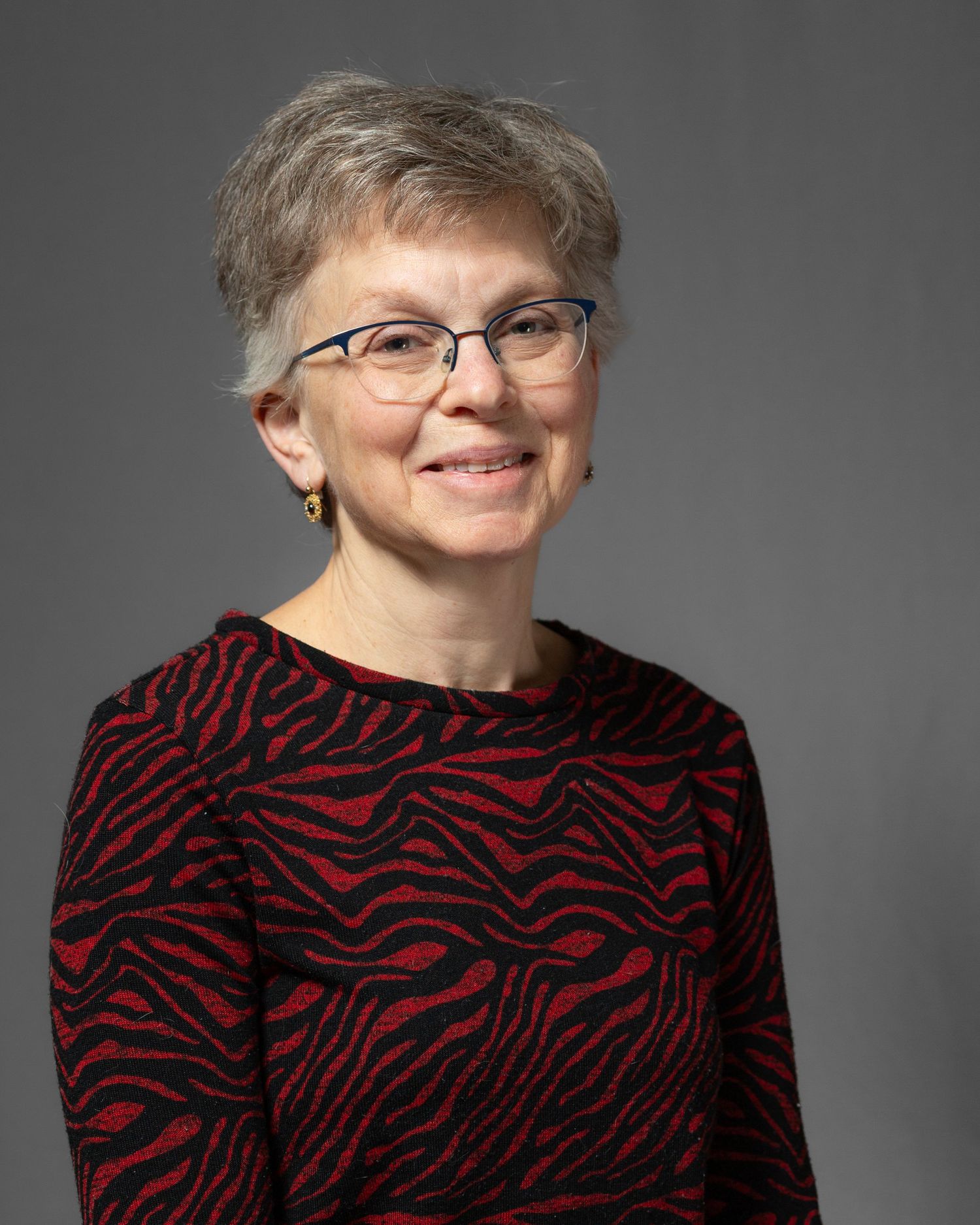 A person in a red and black patterned sweater smiles warmly against a gray studio backdrop.