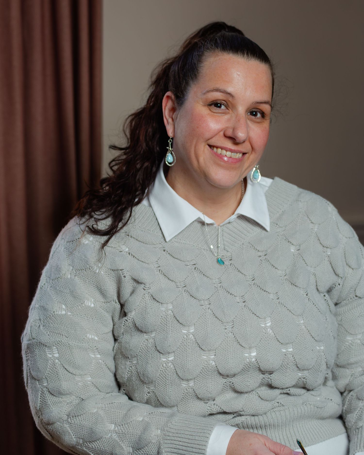 Professional headshot of someone wearing a light gray textured sweater over a white collared shirt with dangling earrings.