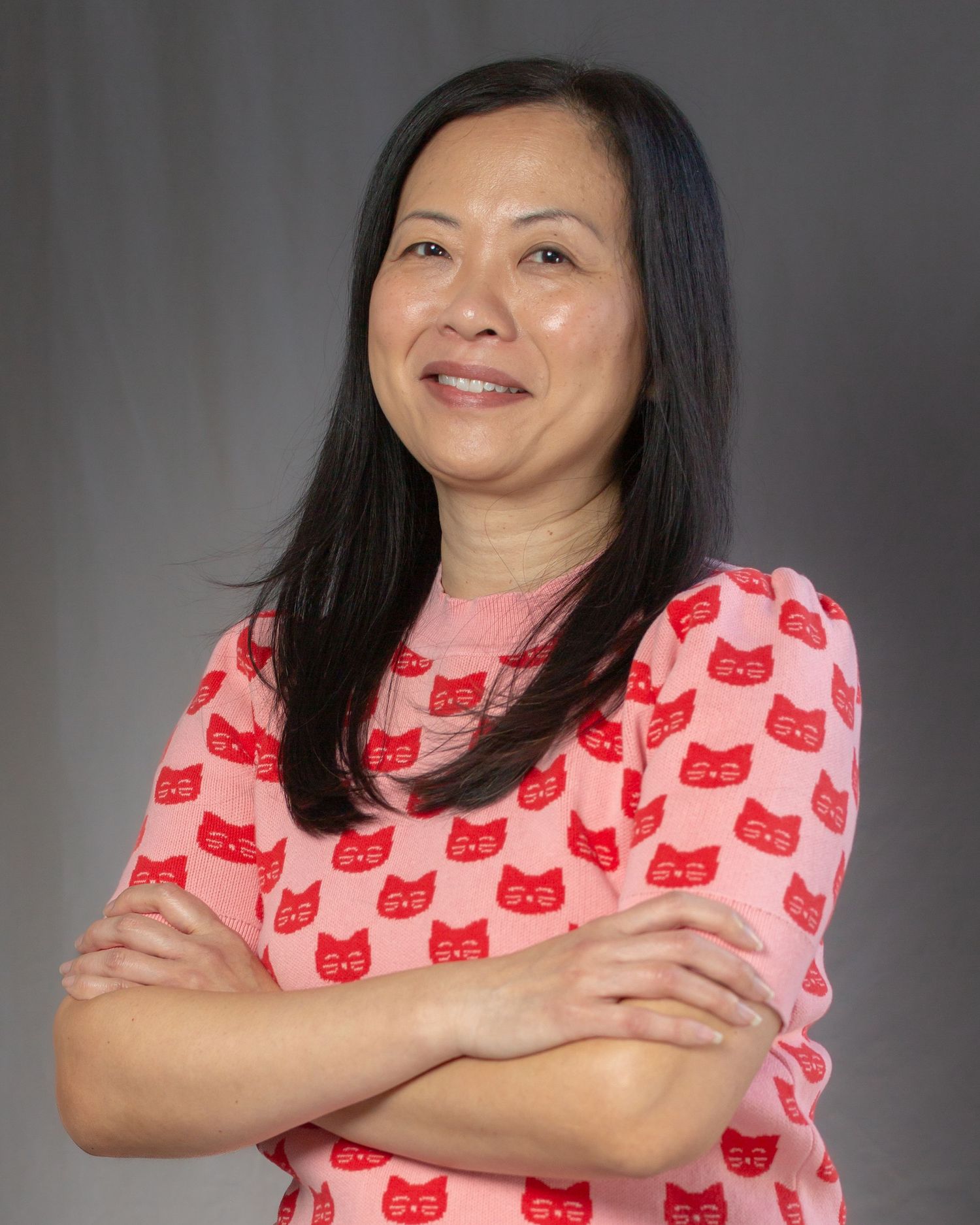 Professional headshot of a person wearing a pink shirt with red cat pattern standing with arms crossed and smiling warmly.