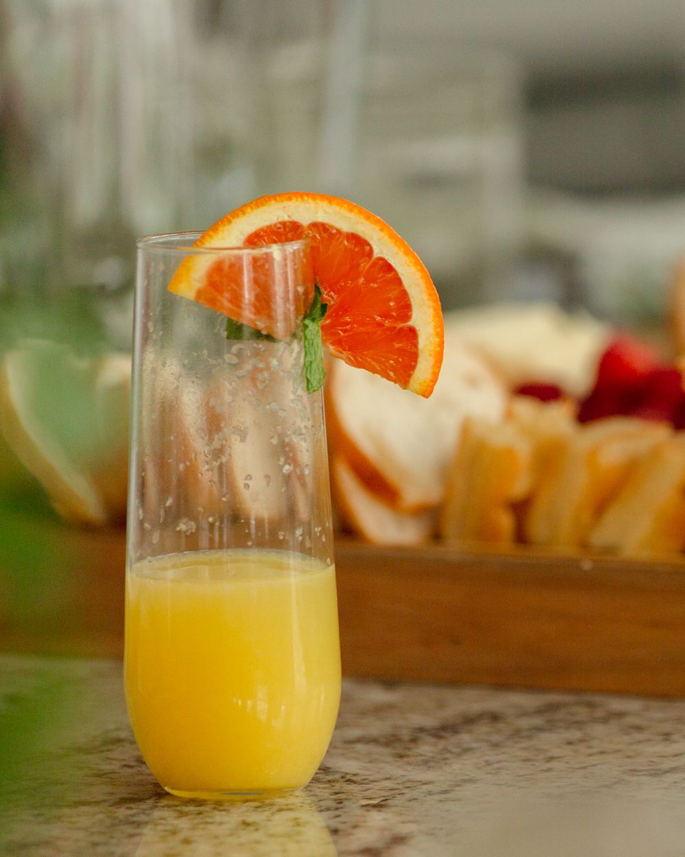 A chilled glass of fresh orange juice garnished with an orange slice and mint, beside a wooden tray of assorted fruits.