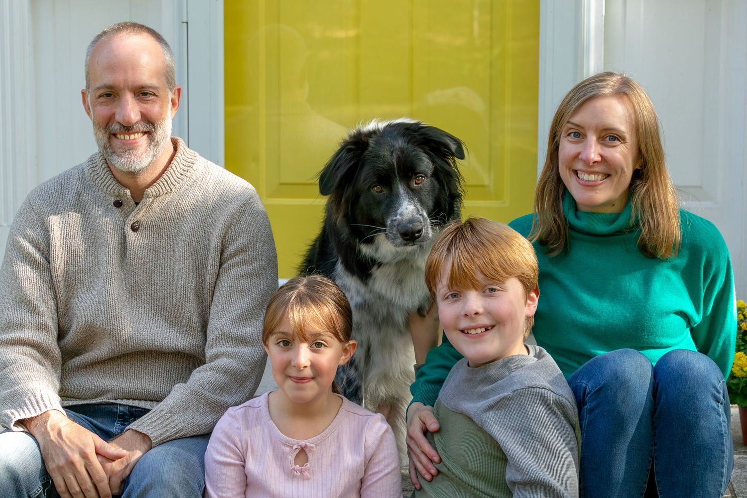 A group of people sit together on a couch wearing casual clothing in front of a yellow wall.