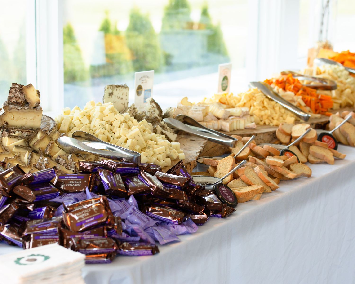 Elegant buffet table with assorted cheeses, bread, snacks, and purple wrapped chocolates at a catered event.