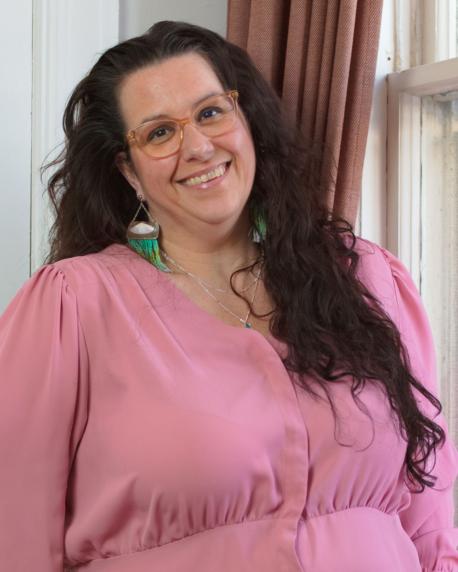 Someone with long dark wavy hair wearing glasses and turquoise earrings smiles warmly while wearing a pink blouse.