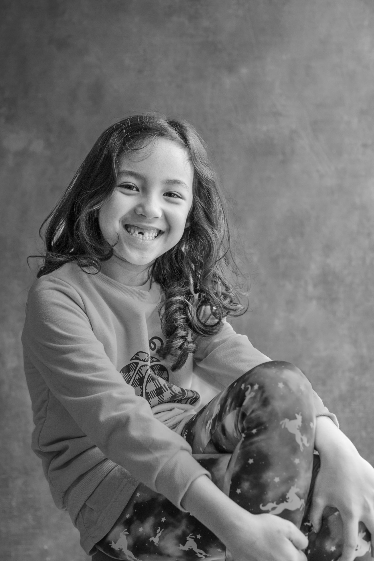 A joyful child in a gray sweater hugs and plays with a plush dinosaur toy against a gray backdrop.