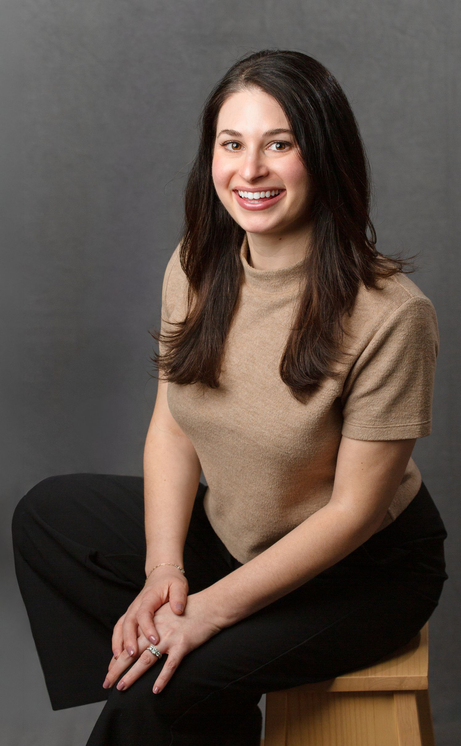 Smiling brunette woman in beige turtleneck and black pants sitting on wooden stool against gray background.