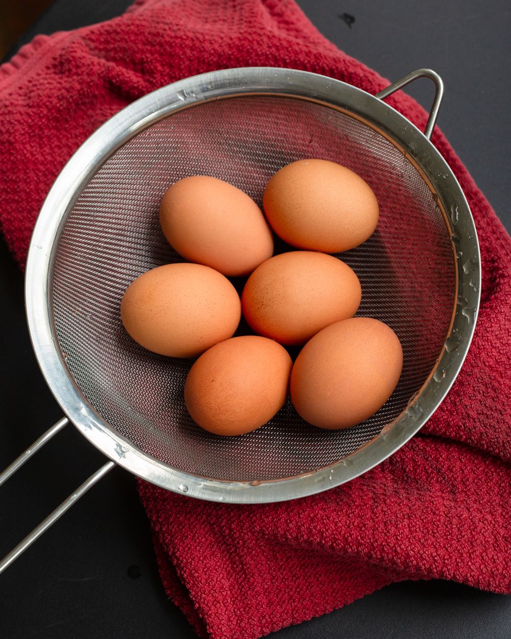 Six fresh brown eggs arranged in a metal mesh colander placed on a red cloth napkin.