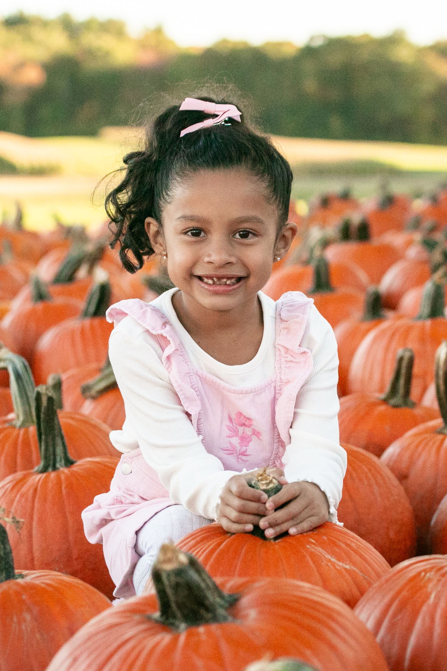 A child in a pink and white dress sits among bright orange pumpkins at a pumpkin patch during autumn.