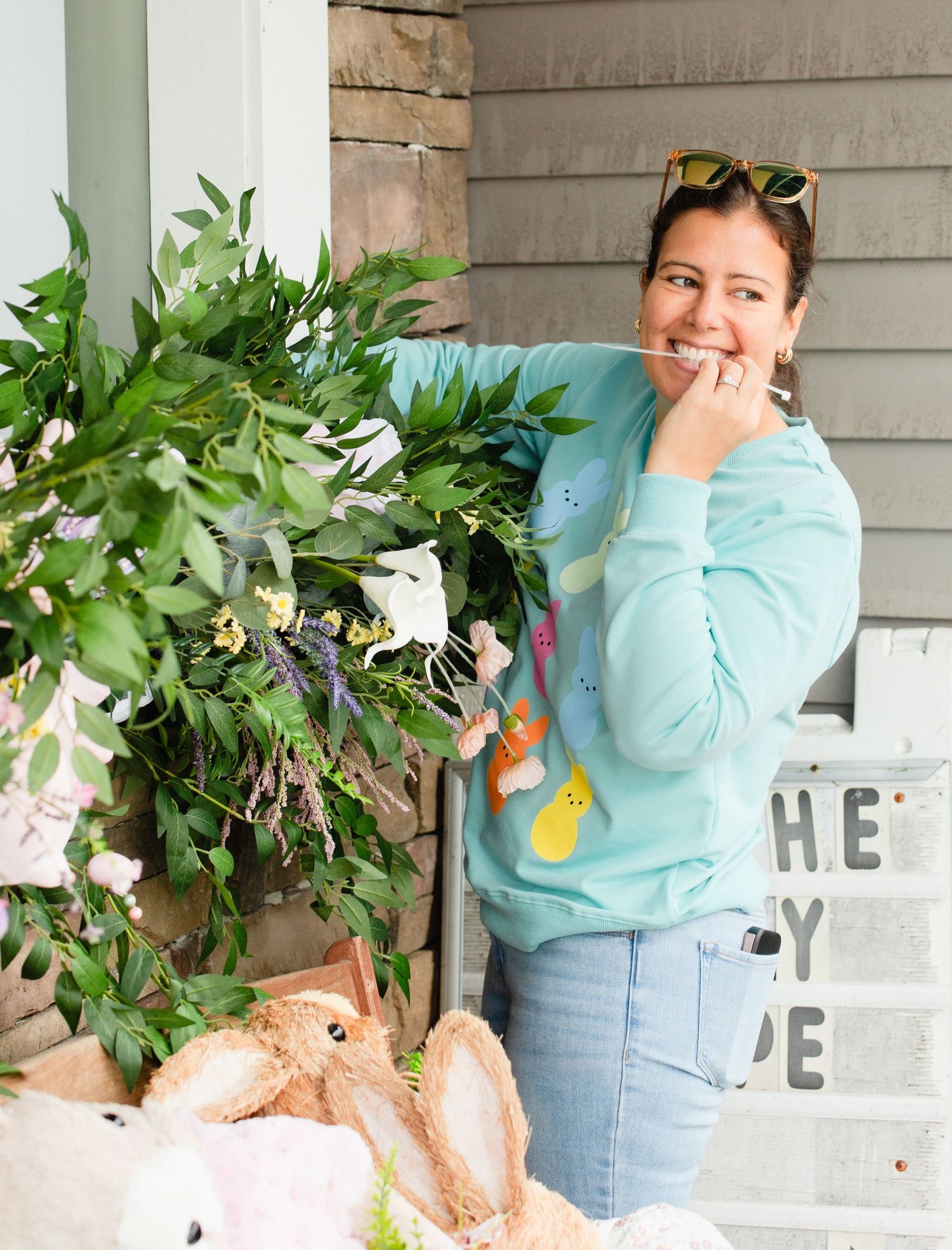 Person in light blue sweater and jeans poses playfully with decorative greenery garland in a home setting.