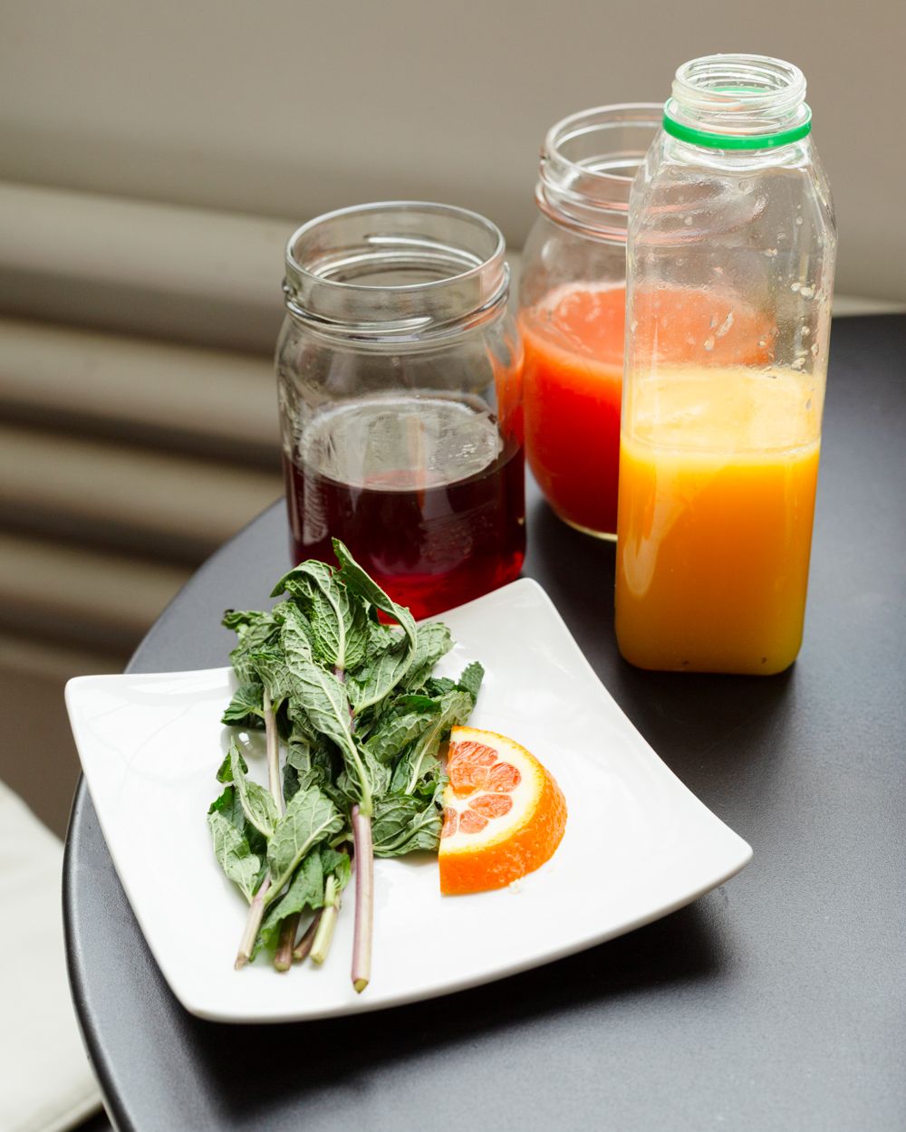 Fresh mint leaves and orange slice on white plate beside jars of colorful juices on dark table.