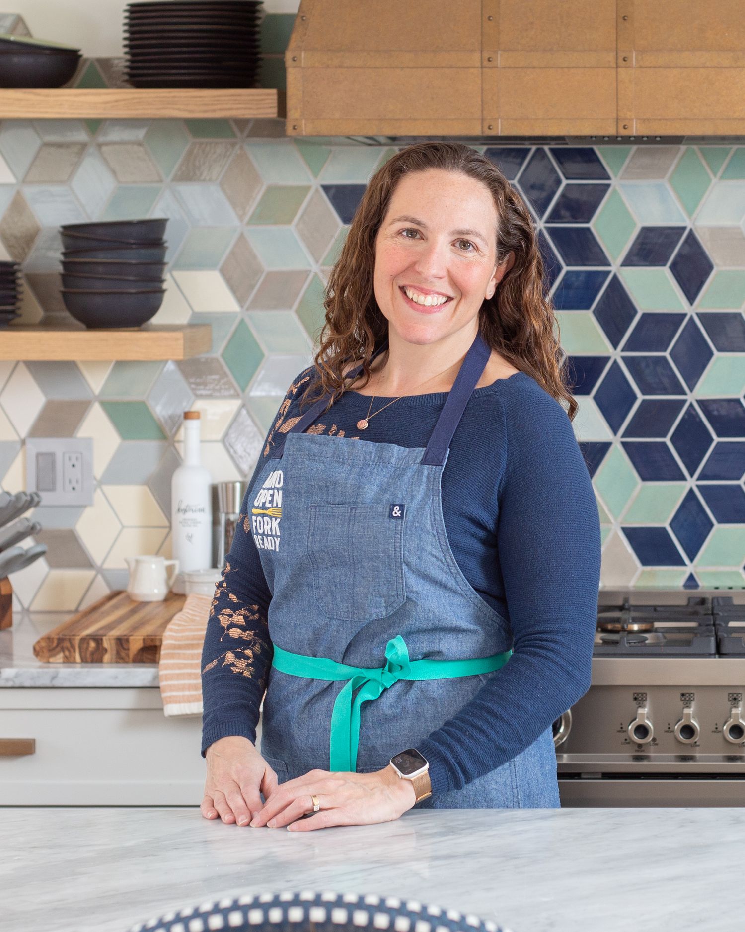 A person wearing a denim apron and navy shirt stands smiling in a modern kitchen with geometric tile backsplash and open shelving.