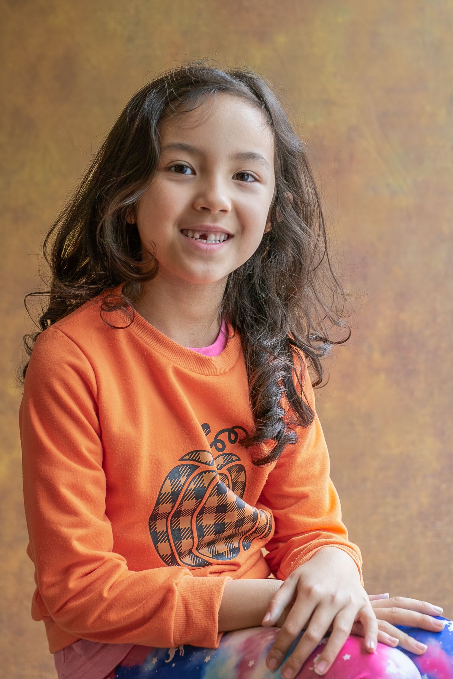 A young child in an orange sweater smiles and plays while sitting against a textured brown background.