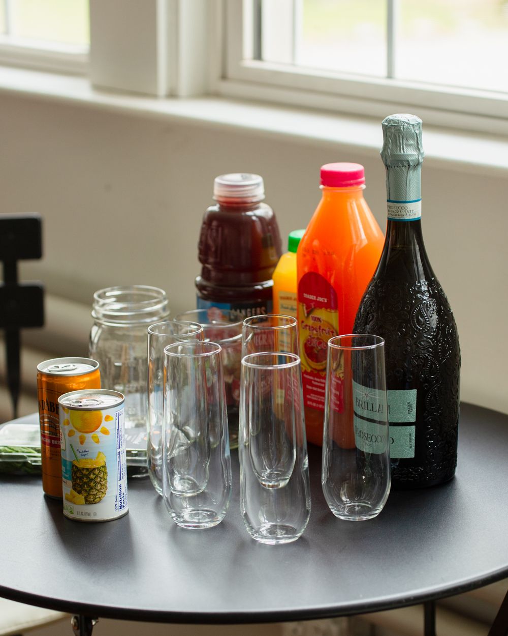 A table with champagne, juice bottles, canned drinks, and clear glasses for a mimosa bar setup.