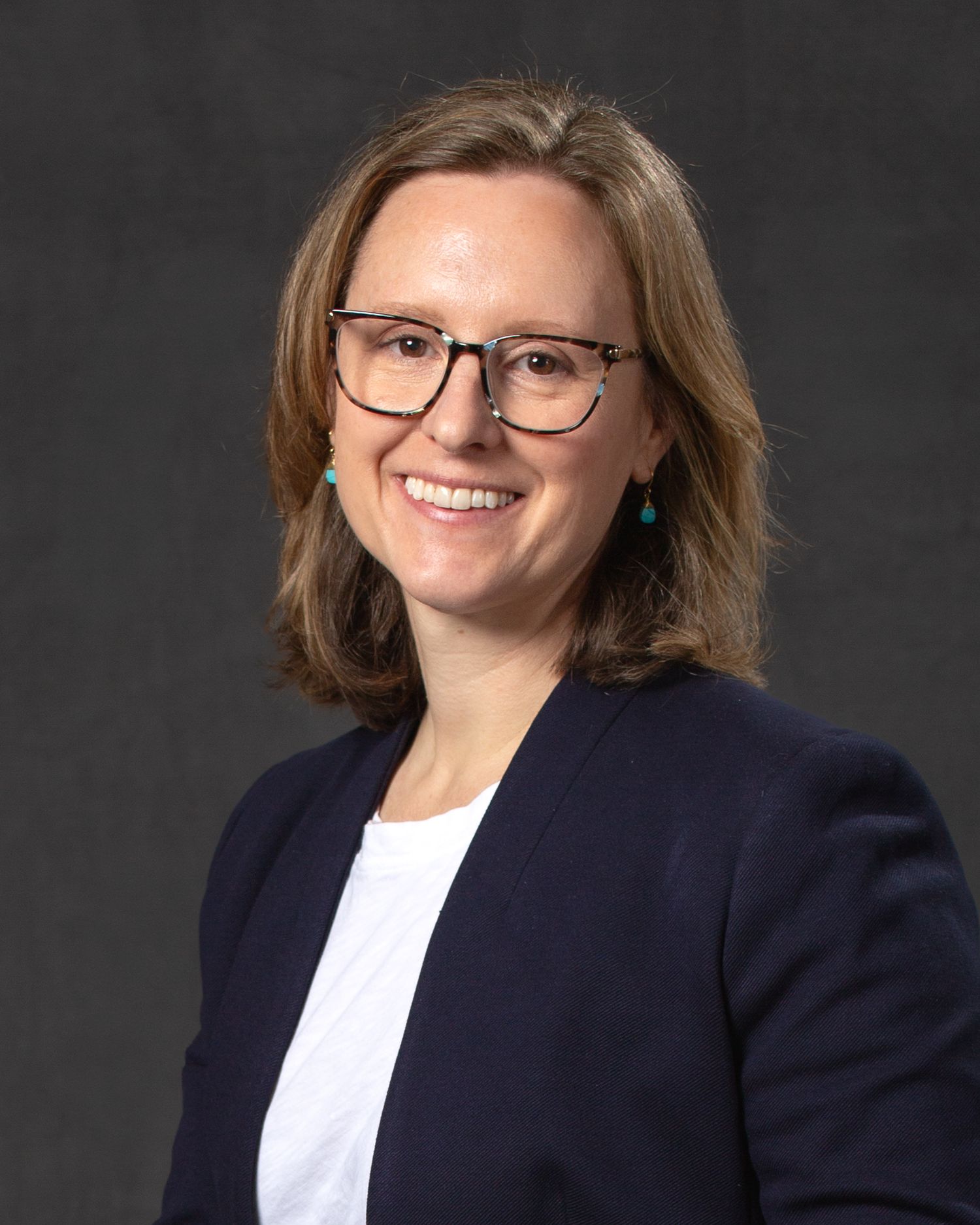 Professional headshot of a smiling woman with glasses and brown hair wearing a navy blazer and teal earrings.
