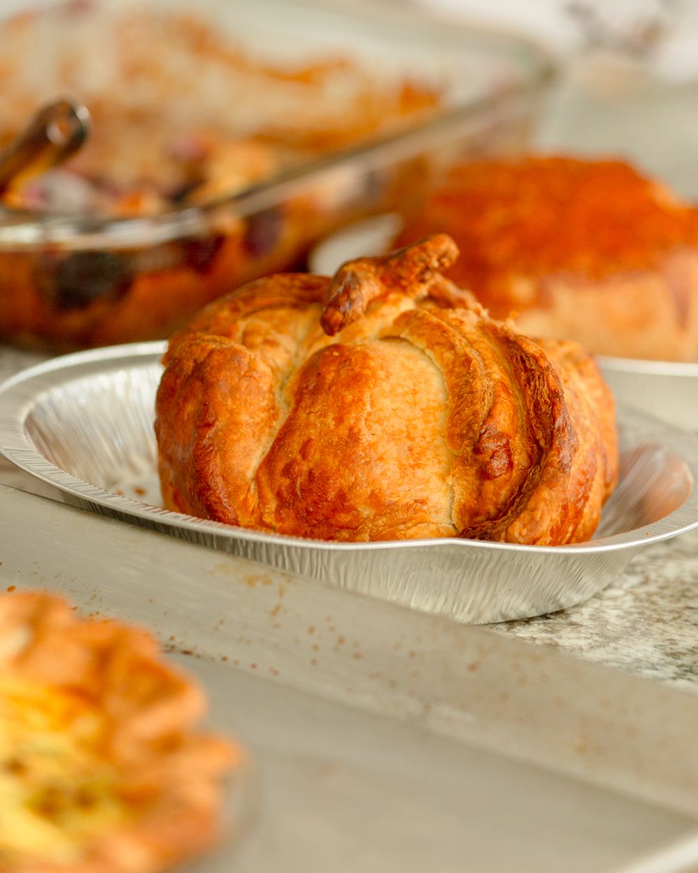 A golden-brown pumpkin-shaped pastry bun in a foil dish, with a baked casserole dish visible in the background.