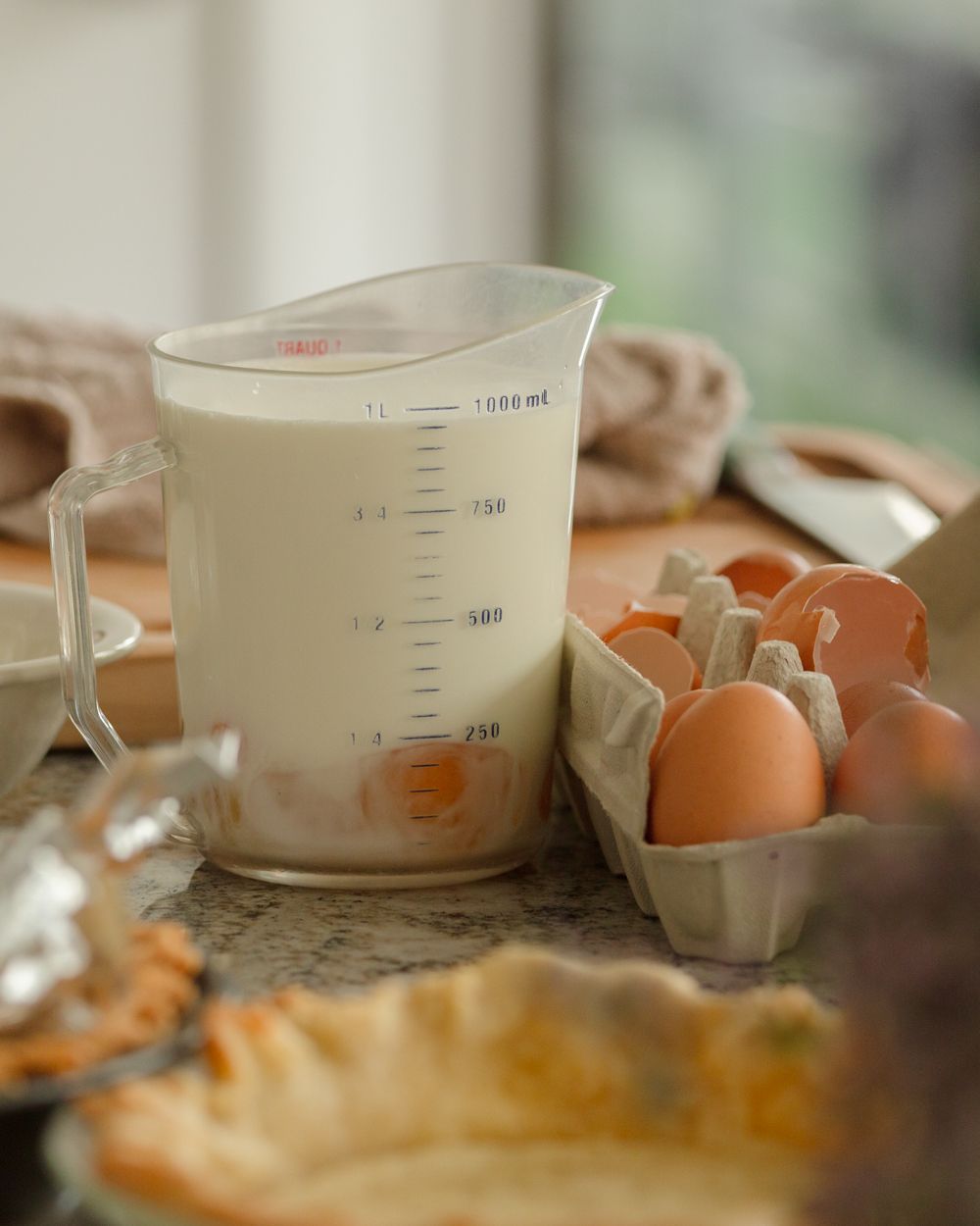 Large measuring cup filled with milk beside brown eggs in carton on a kitchen counter.
