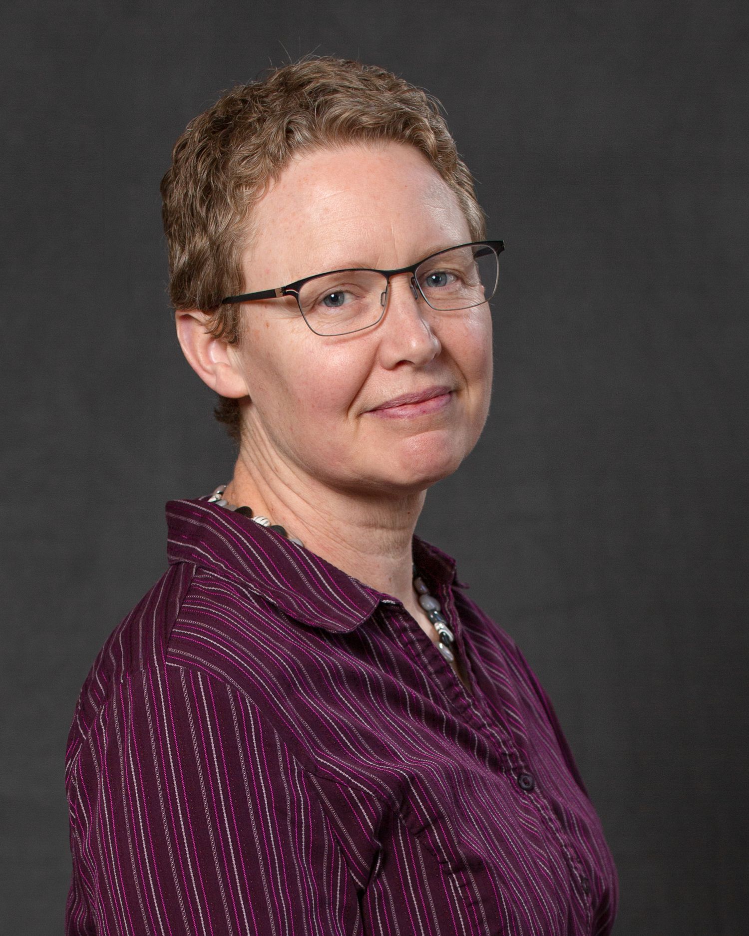 Woman with short hair and glasses wearing purple striped shirt in professional portrait photo.