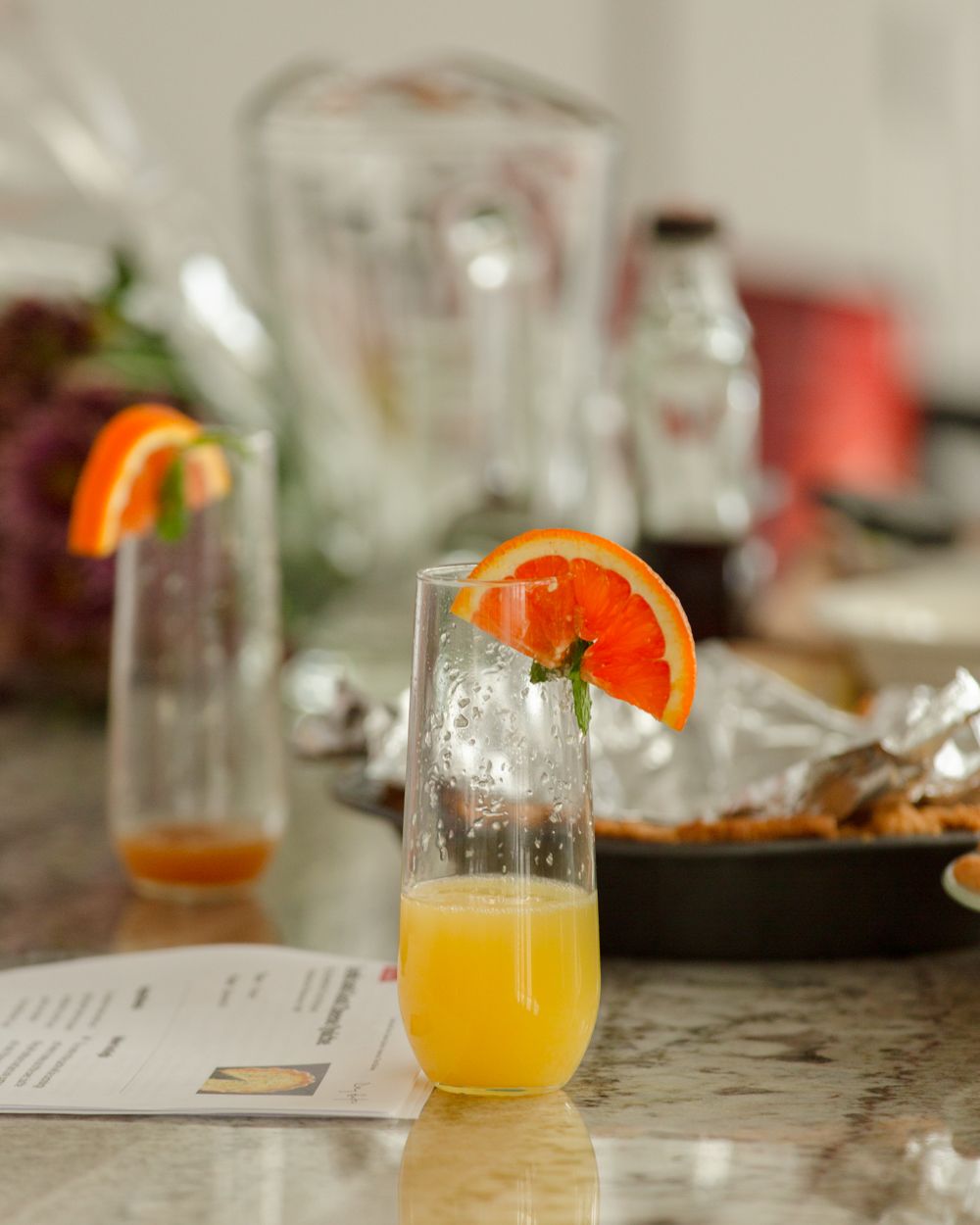 Two stemless champagne glasses with orange juice and citrus garnish on a marble countertop at a brunch spread.