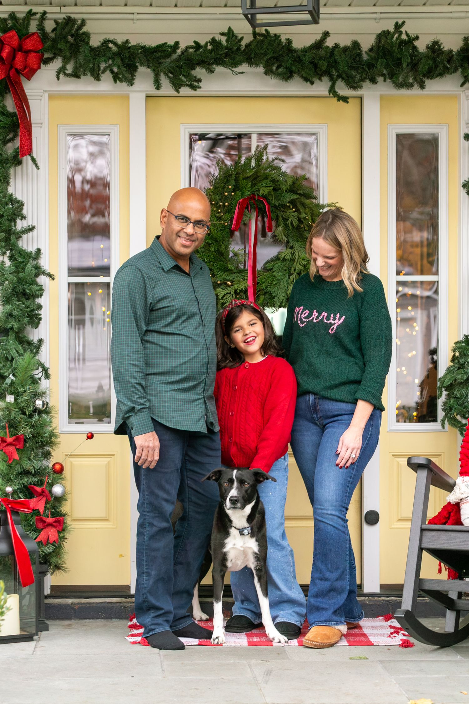 Family poses on festive front porch decorated with Christmas garland and red bows while two small dogs sit nearby.