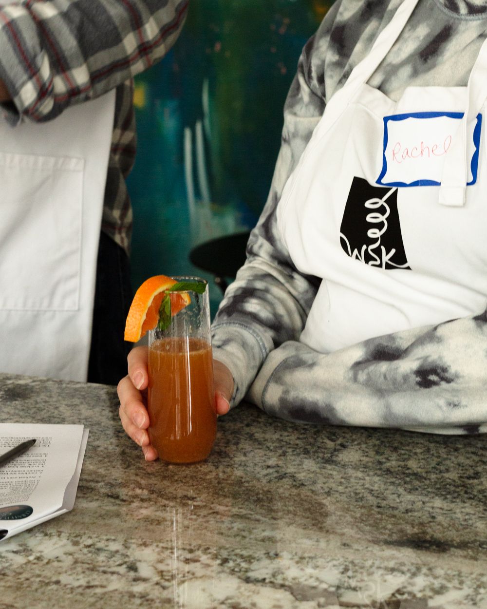 Person in tie-dye shirt and white apron holding a fresh orange garnished cocktail at a counter.