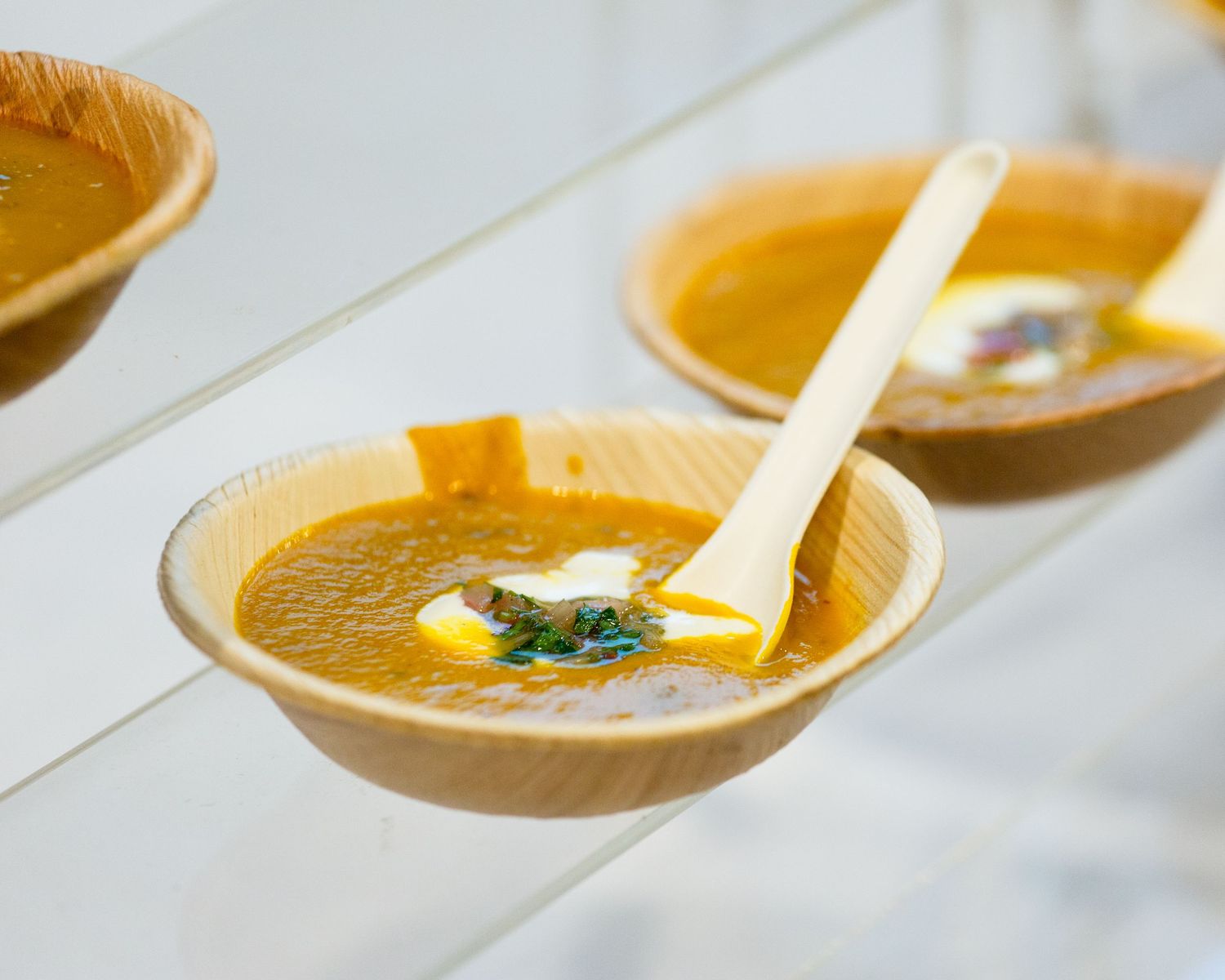 Three wooden bowls of golden soup garnished with cream and herbs, served with white spoons on a white surface.