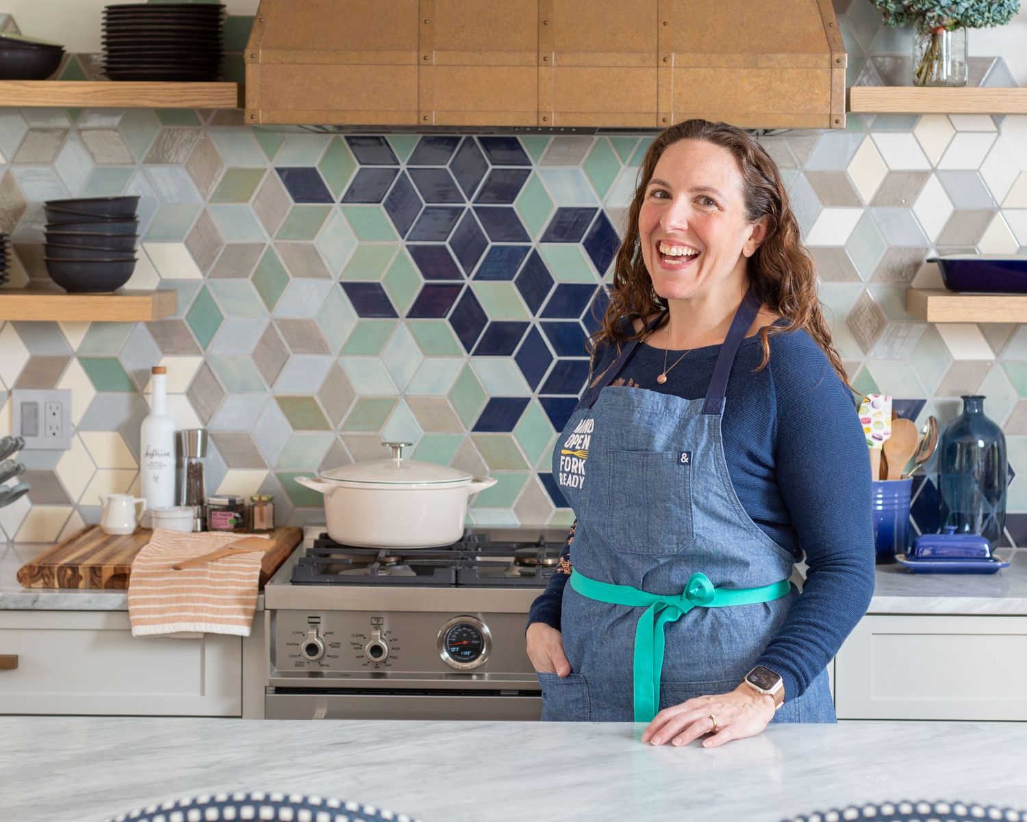 A person in a blue top demonstrates cooking in a modern kitchen with geometric tile backsplash and wooden shelving.
