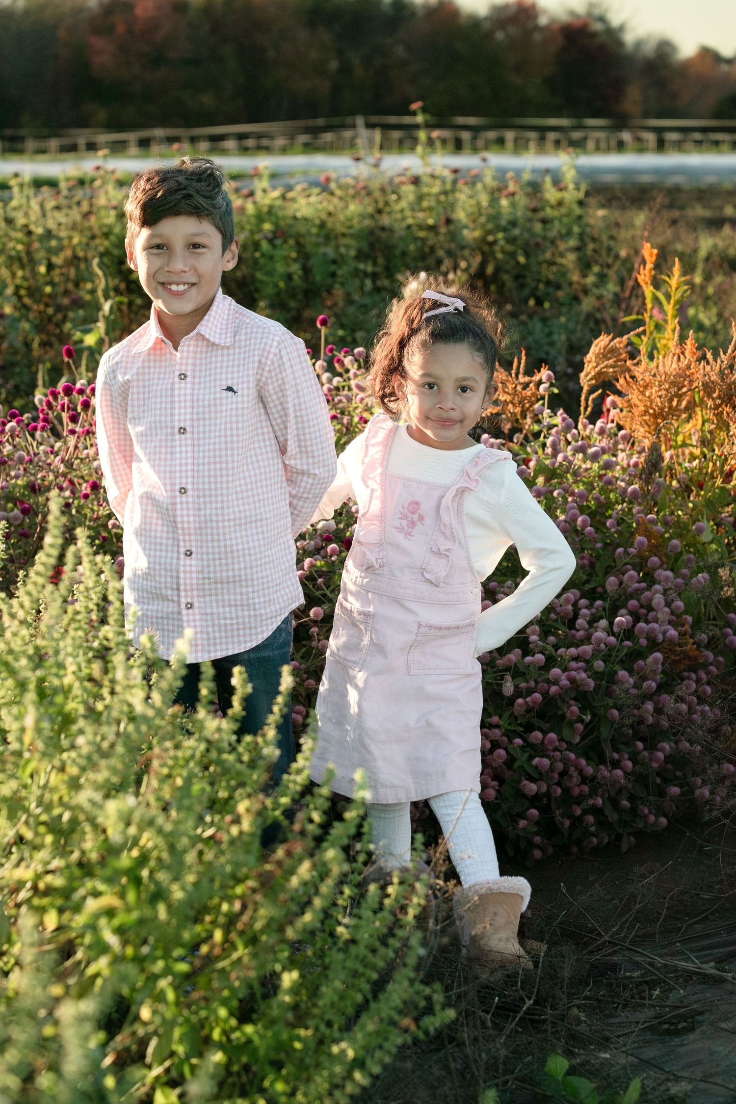 Children pose together in a garden at sunset among colorful flowers and greenery wearing coordinated white outfits.