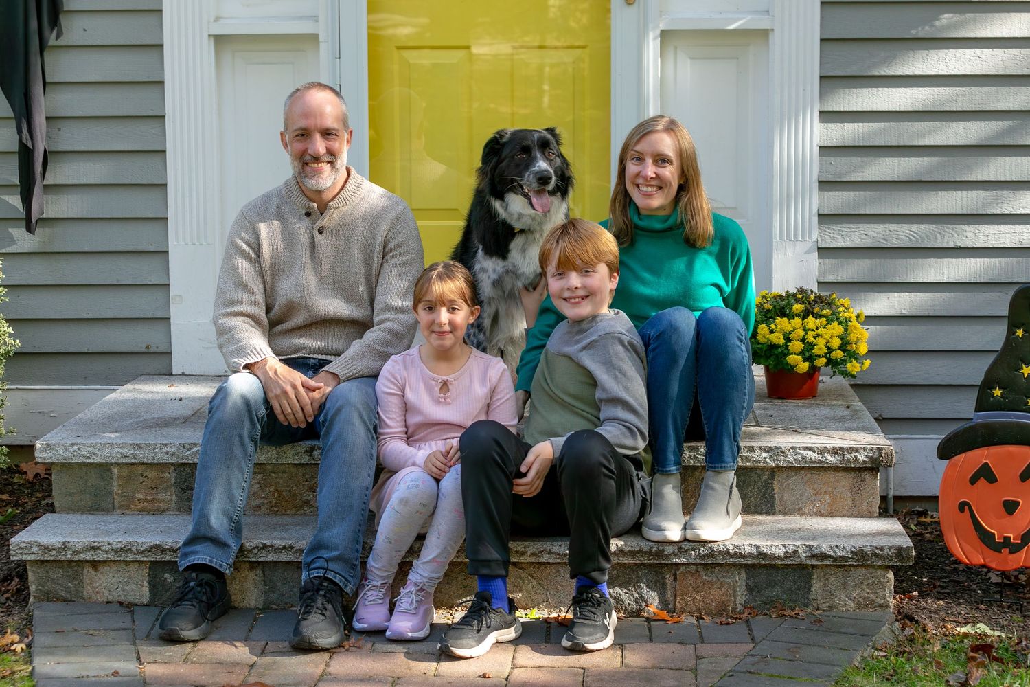 A family sits together on the front steps of their house with fall decorations visible.